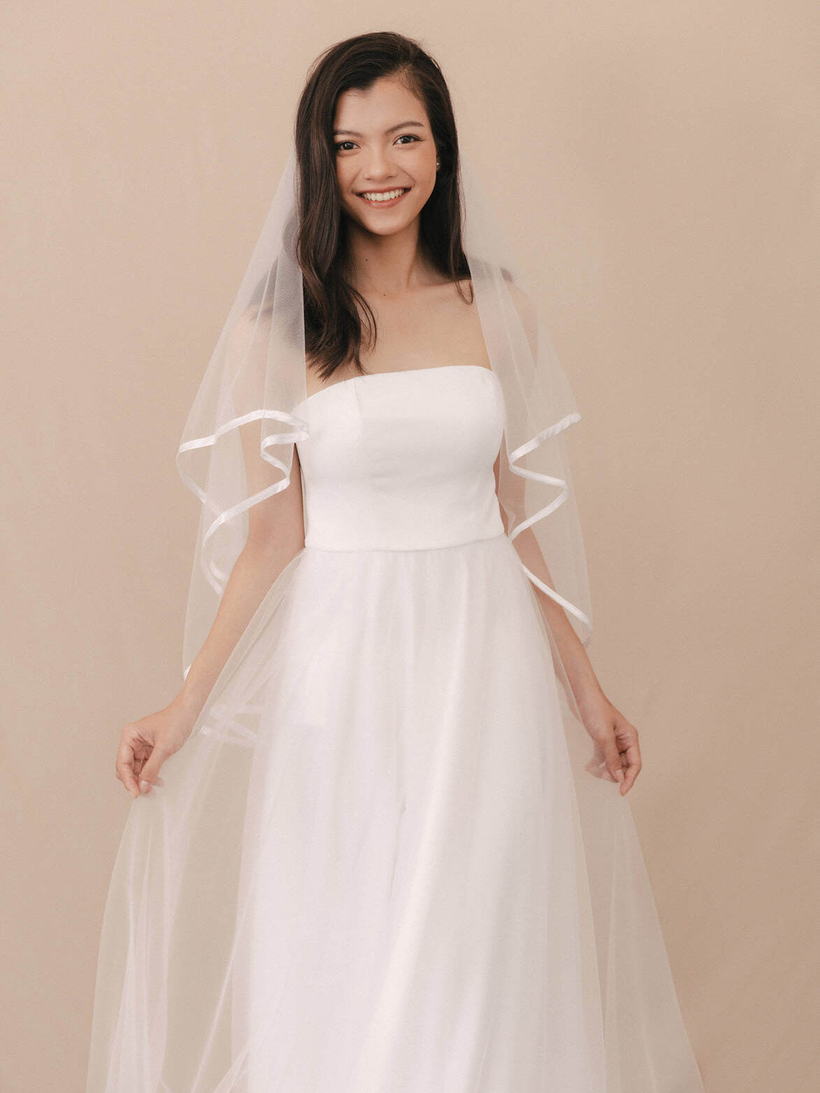 Woman wearing a strapless white wedding dress with a sheer overlay on a beige background