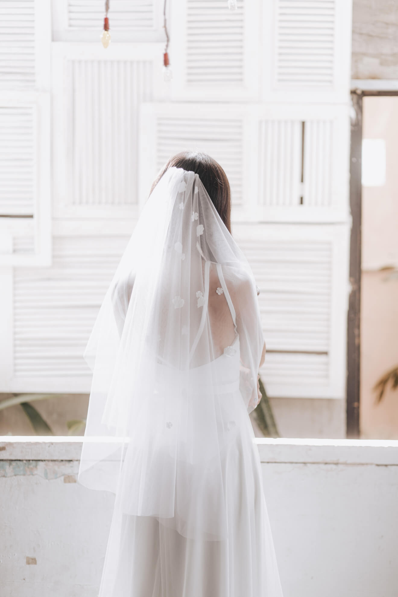 Person wearing a white wedding dress with a veil in front of a window with blinds.