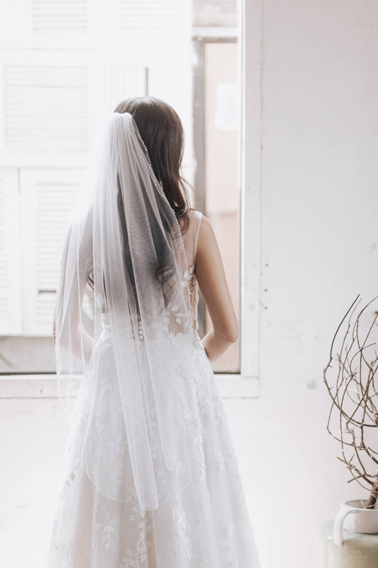 Woman in a wedding dress with a veil standing in front of a window.