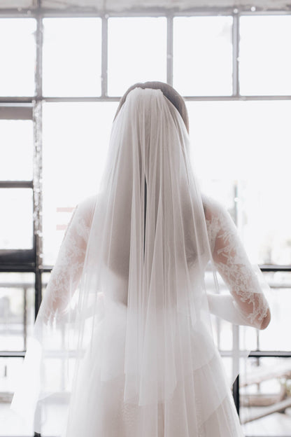 Person wearing a long white wedding dress with a veil in front of a window.