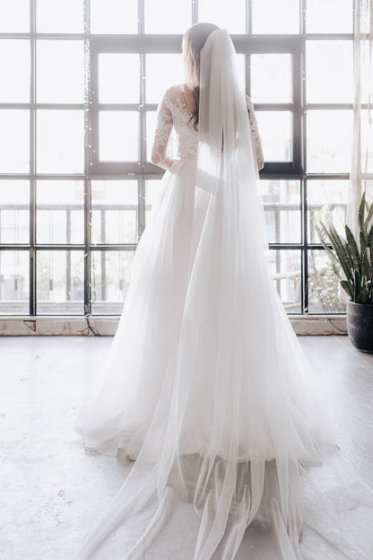 Person wearing a white wedding dress with a long veil in a bright room with large windows.
