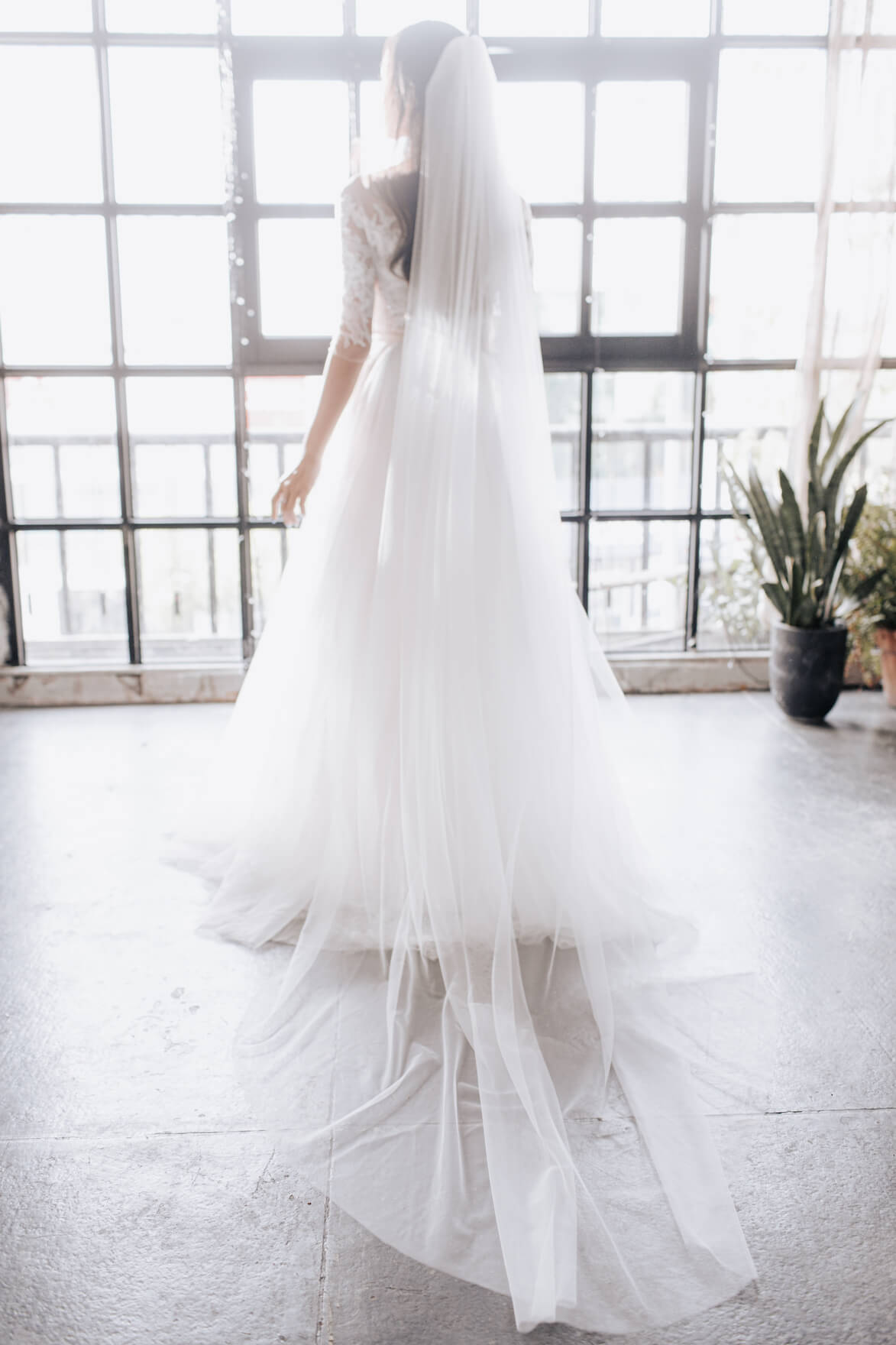 Person wearing a long white wedding dress with a veil in a bright room with large windows.