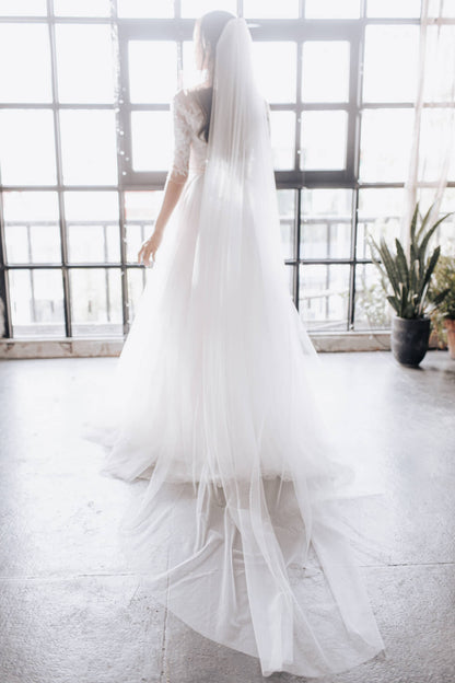 Person wearing a long white wedding dress with a veil in a bright room with large windows.