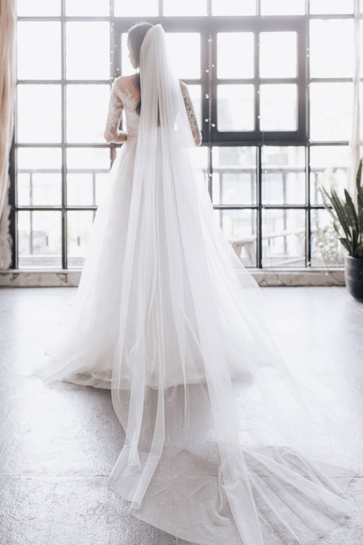 Person wearing a long white wedding dress with a veil in a bright room with large windows.