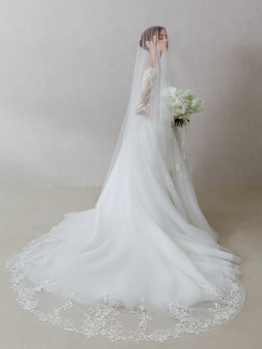 Woman in a white wedding dress with a long veil holding a bouquet against a plain background