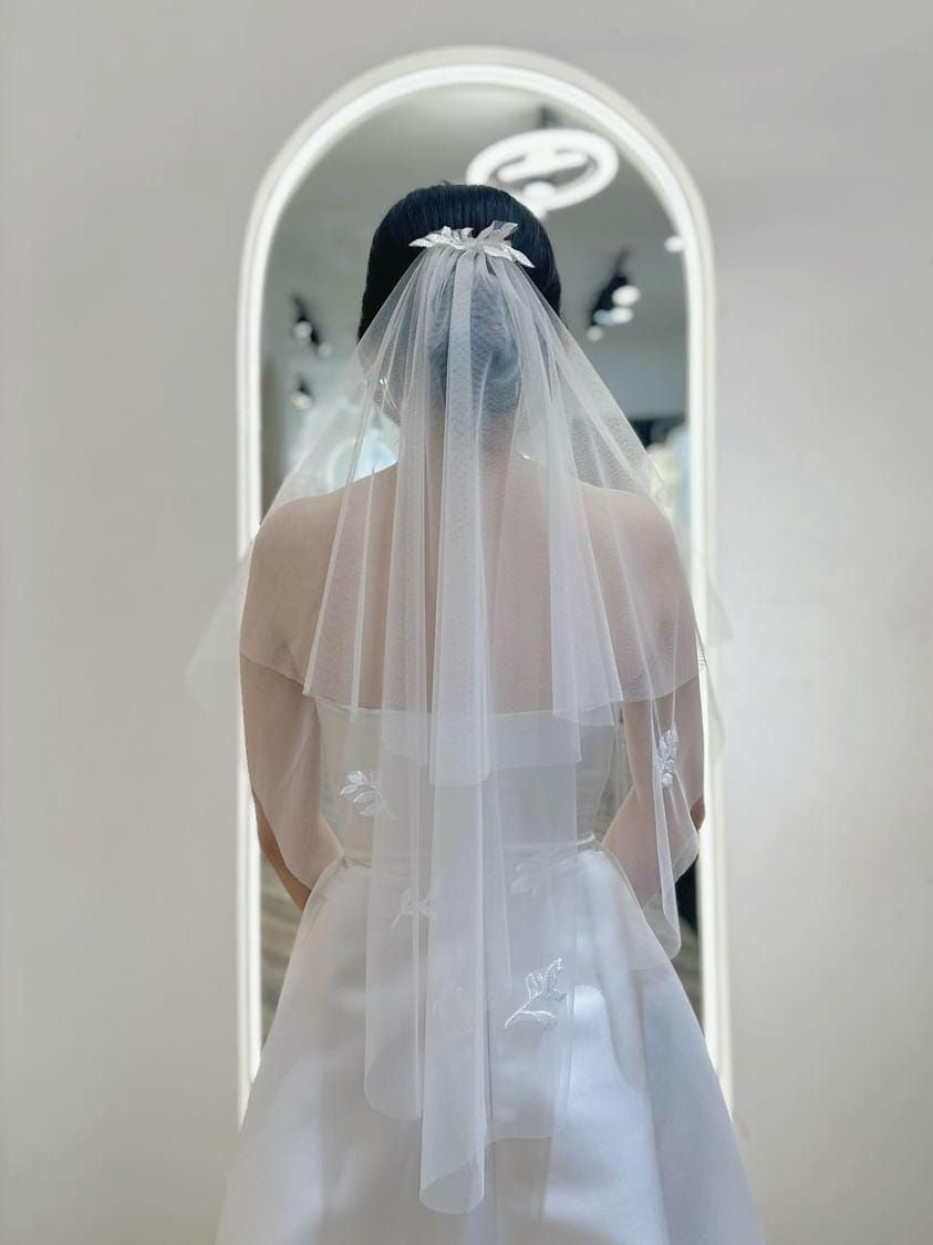 Bride wearing a Graceful Embroidered Veil with floral appliqué and ribbon detail, showcasing the layered tulle design in a bridal boutique.