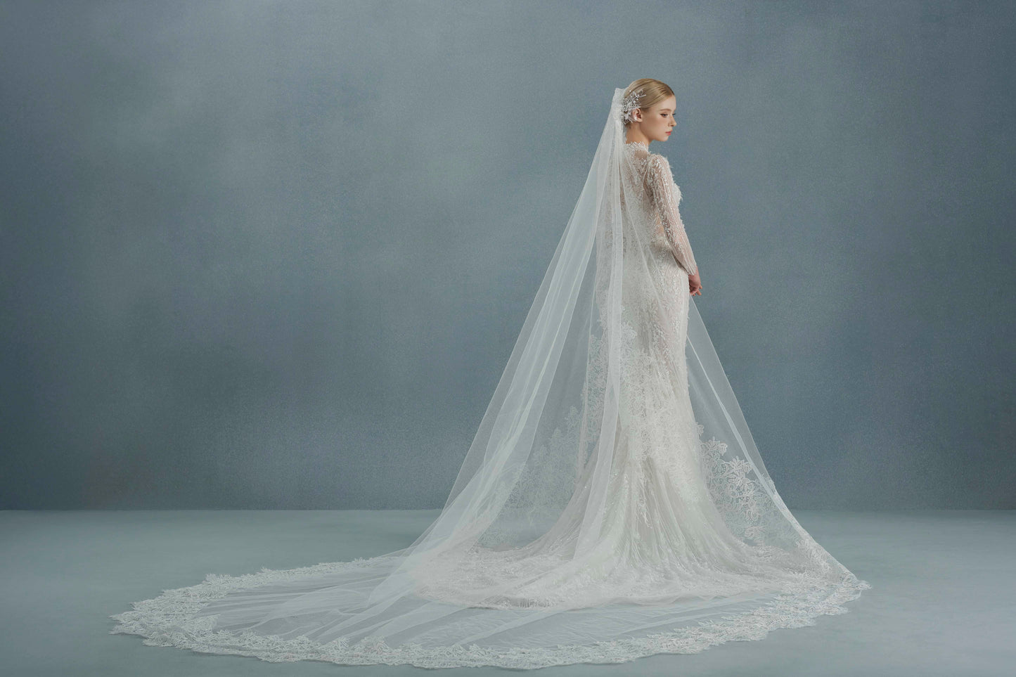Bride in a long-sleeve wedding dress with a cathedral-length veil against a gray background