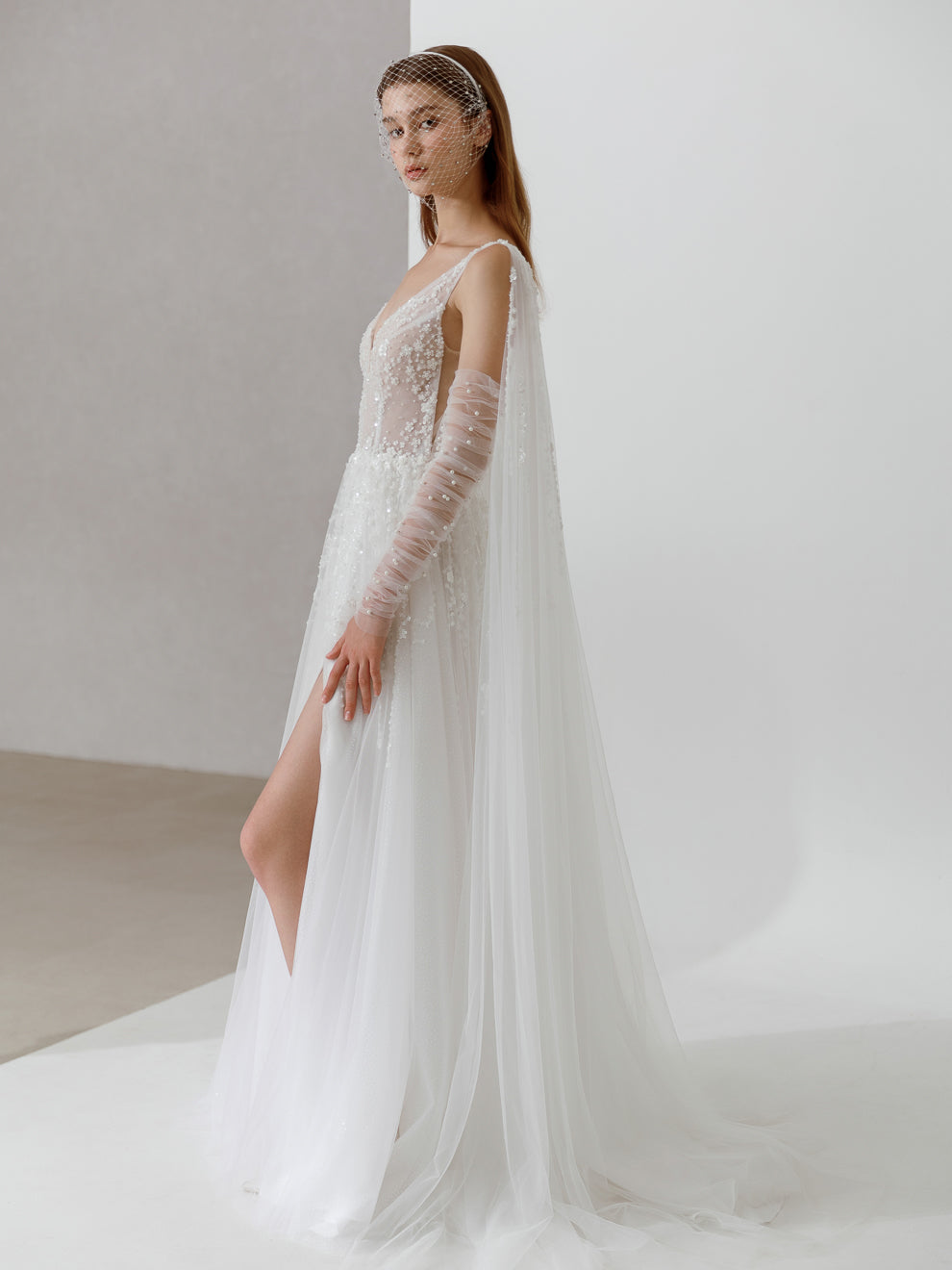 Model wearing a white wedding dress with long sleeves and a slit on a plain background