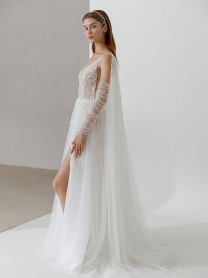 Model wearing a white wedding dress with long sleeves and a slit on a plain background