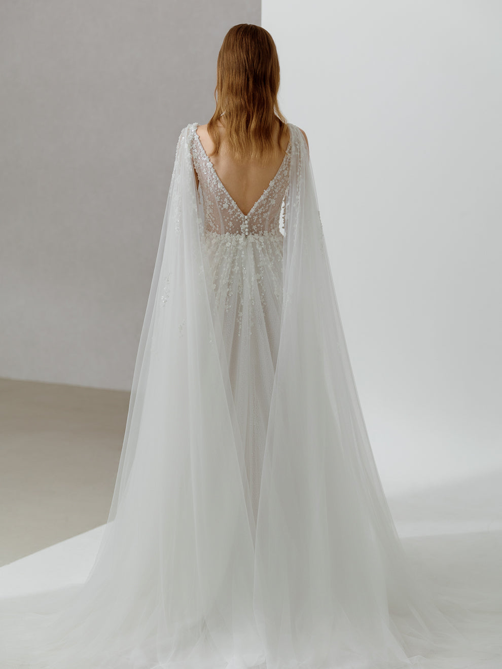Woman wearing a white wedding dress with a long train and beaded back, standing against a plain background.