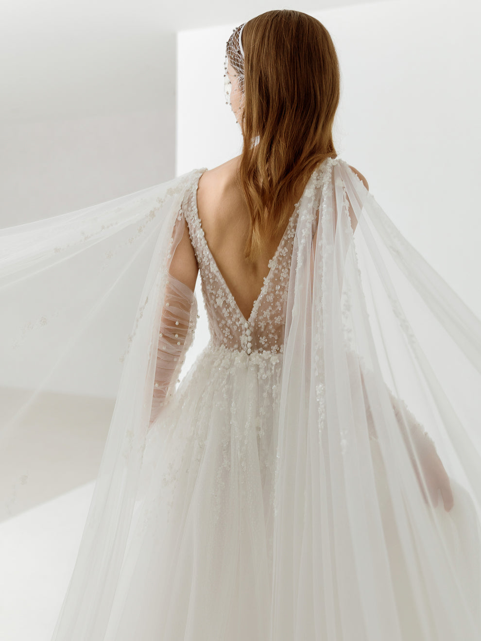 Woman in a white wedding dress with long sleeves and a sheer overlay, standing against a white background.