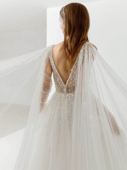 Woman in a white wedding dress with long sleeves and a sheer overlay, standing against a white background.