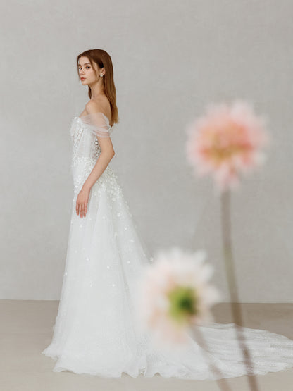 Woman in a white wedding dress standing against a neutral background with flowers in the foreground.