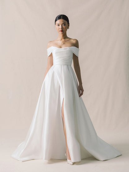 Woman wearing a white off-shoulder gown with a high slit on a plain background