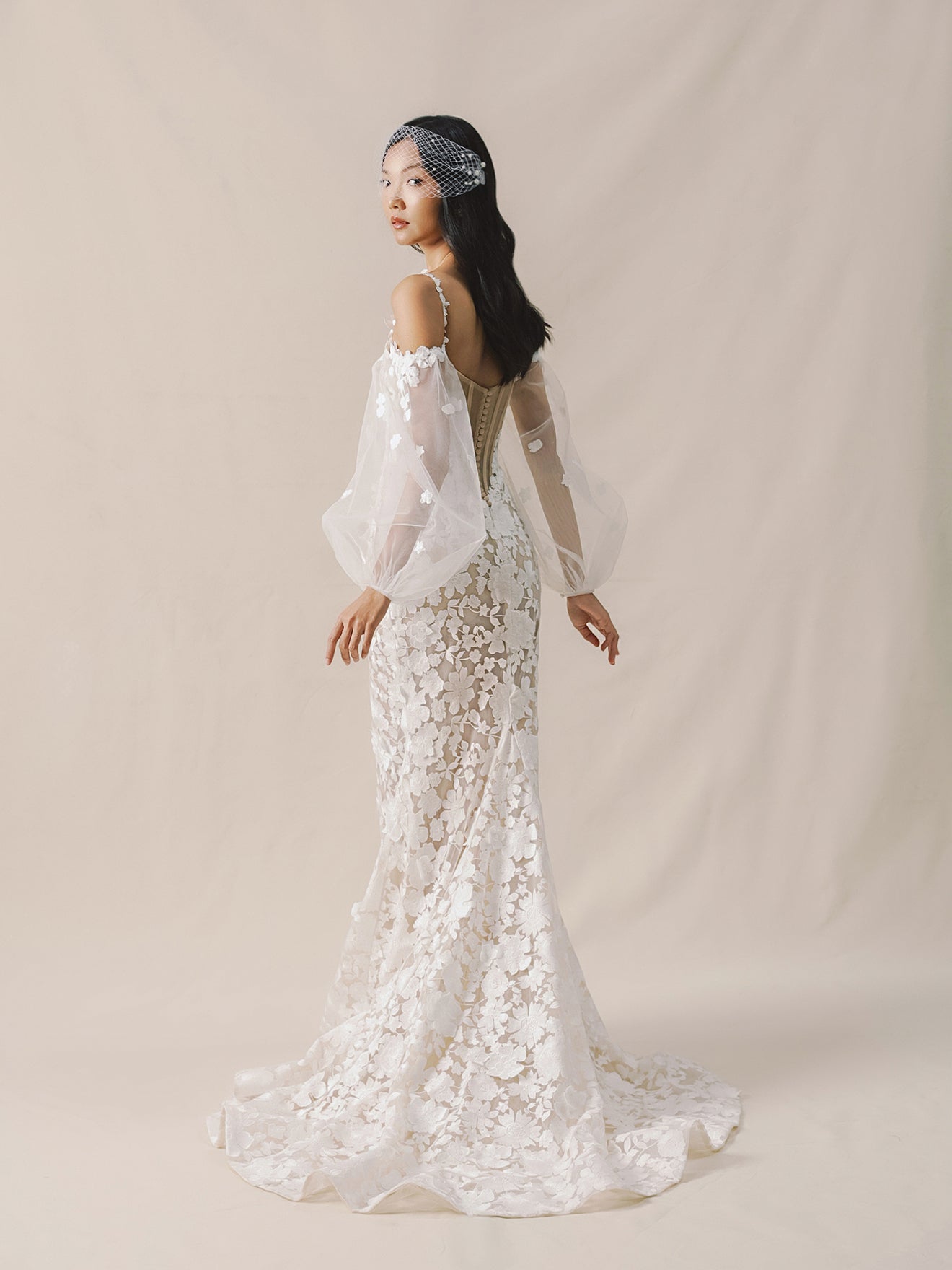 Woman wearing a white lace wedding dress with sheer sleeves against a beige background