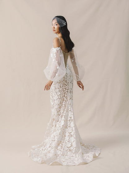 Woman wearing a white lace wedding dress with sheer sleeves against a beige background