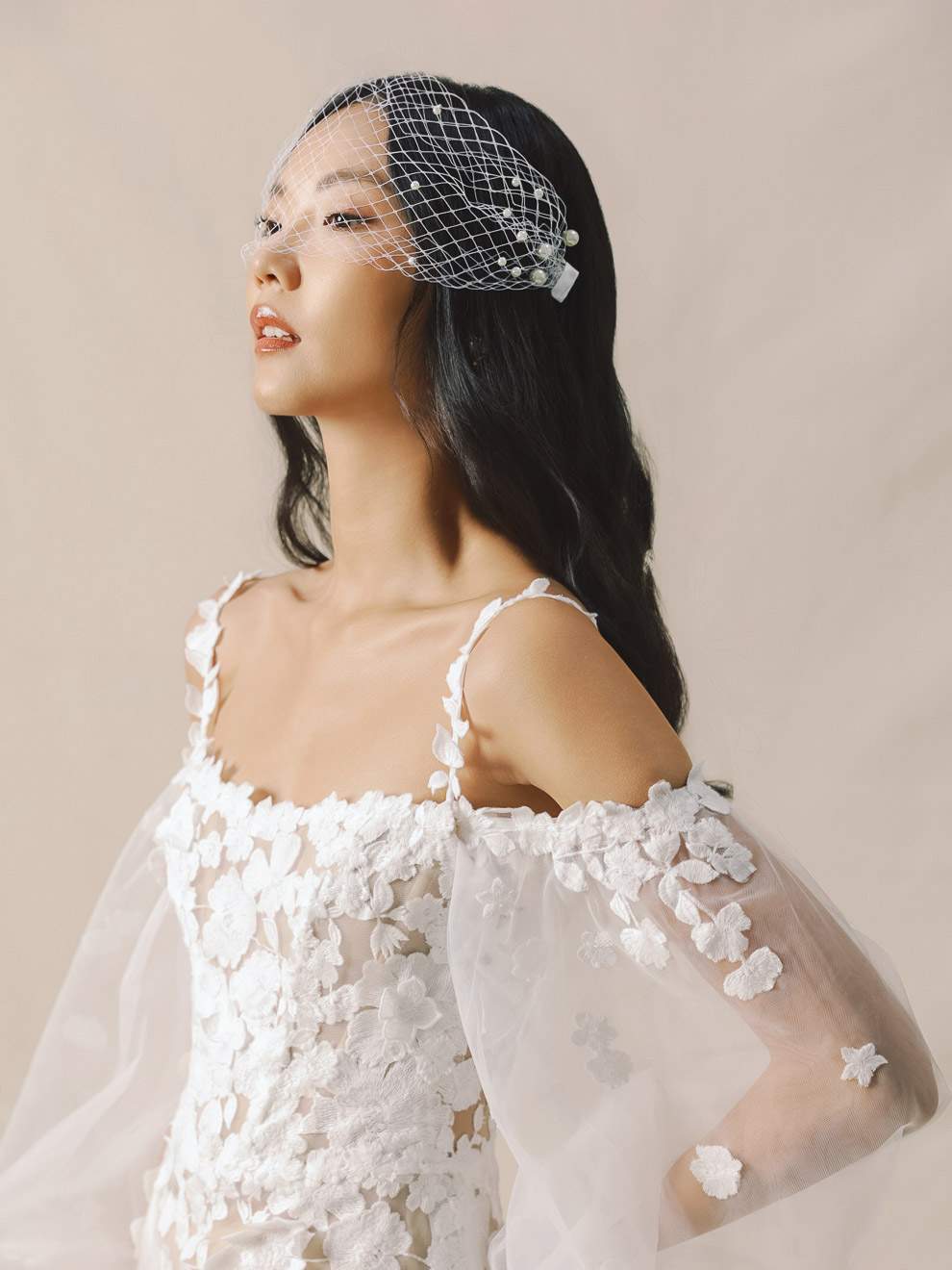 Woman wearing a white lace dress with floral details and a birdcage veil against a beige background