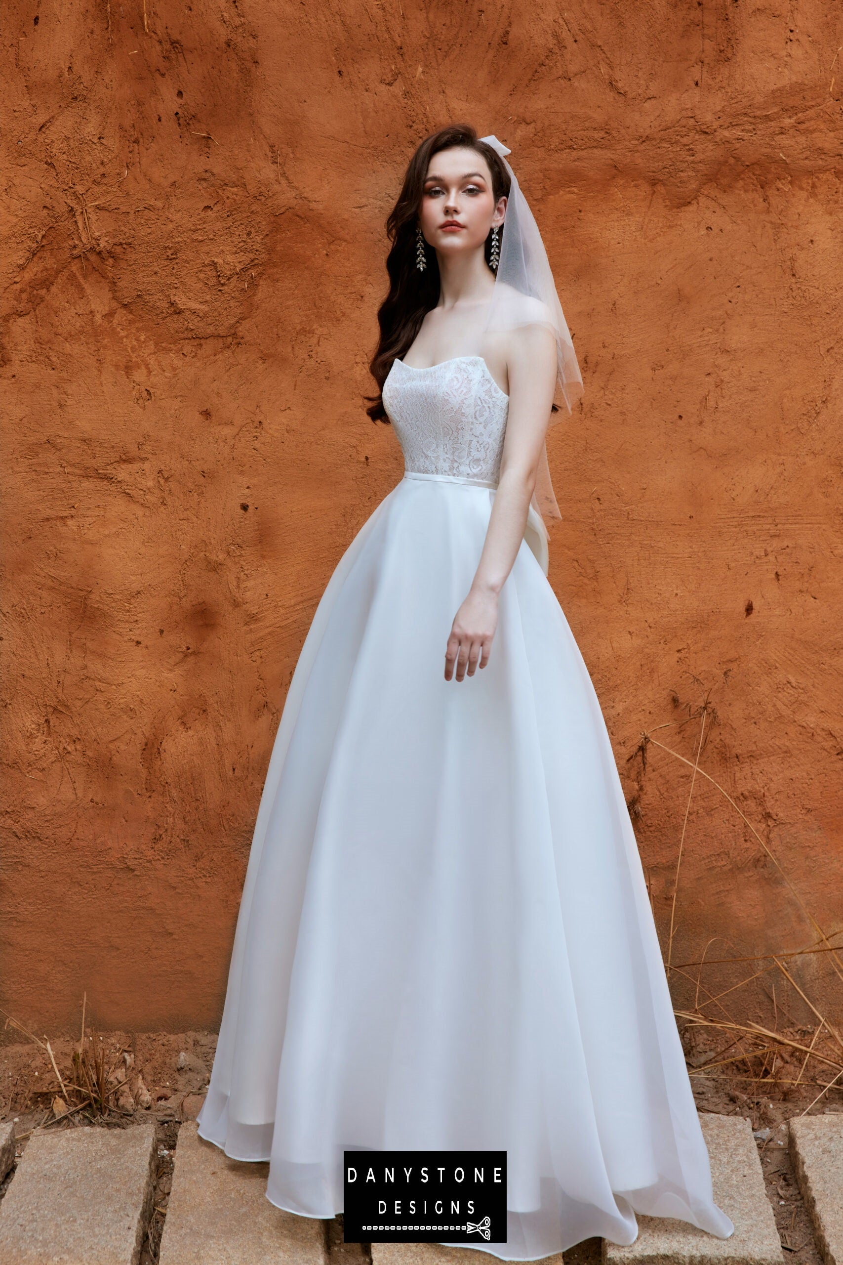 Side view of the model in the 2-in-1 wedding dress with a lace bodice and silk chiffon skirt, standing against a warm, textured background.