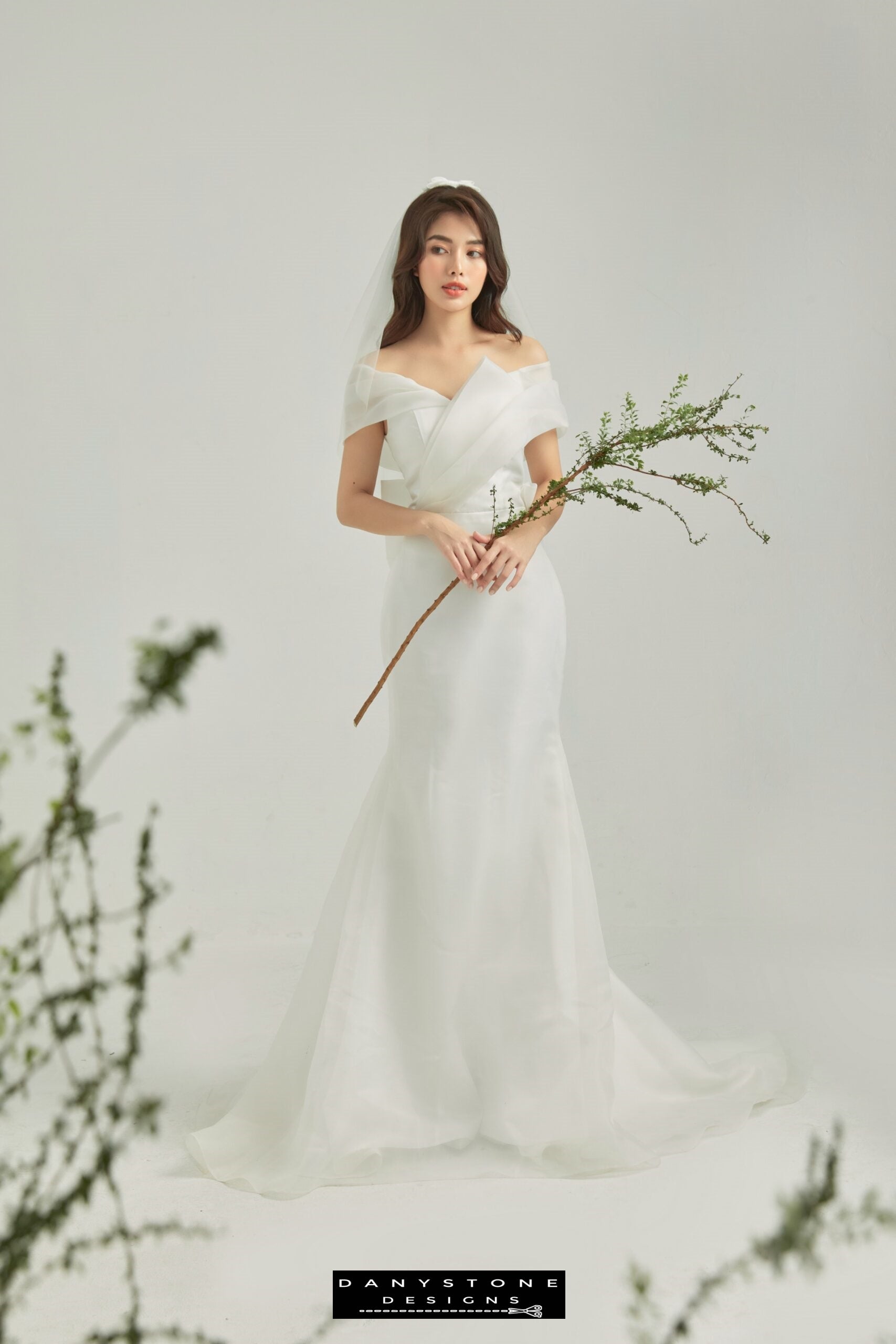 Bride wearing a modern off-shoulder wedding gown with wrap detail, holding a branch – Danystone Designs