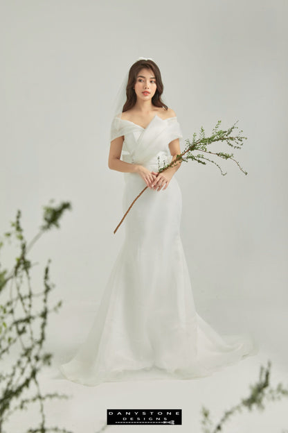 Bride wearing a modern off-shoulder wedding gown with wrap detail, holding a branch – Danystone Designs