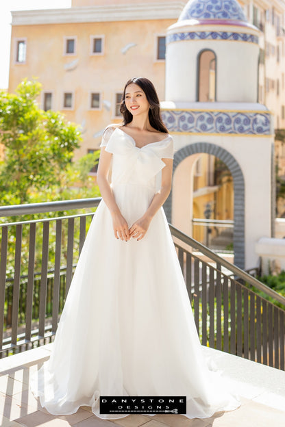 Image 4: Full-length view of a bride in a silk chiffon wedding dress with bow accents, standing on a balcony.