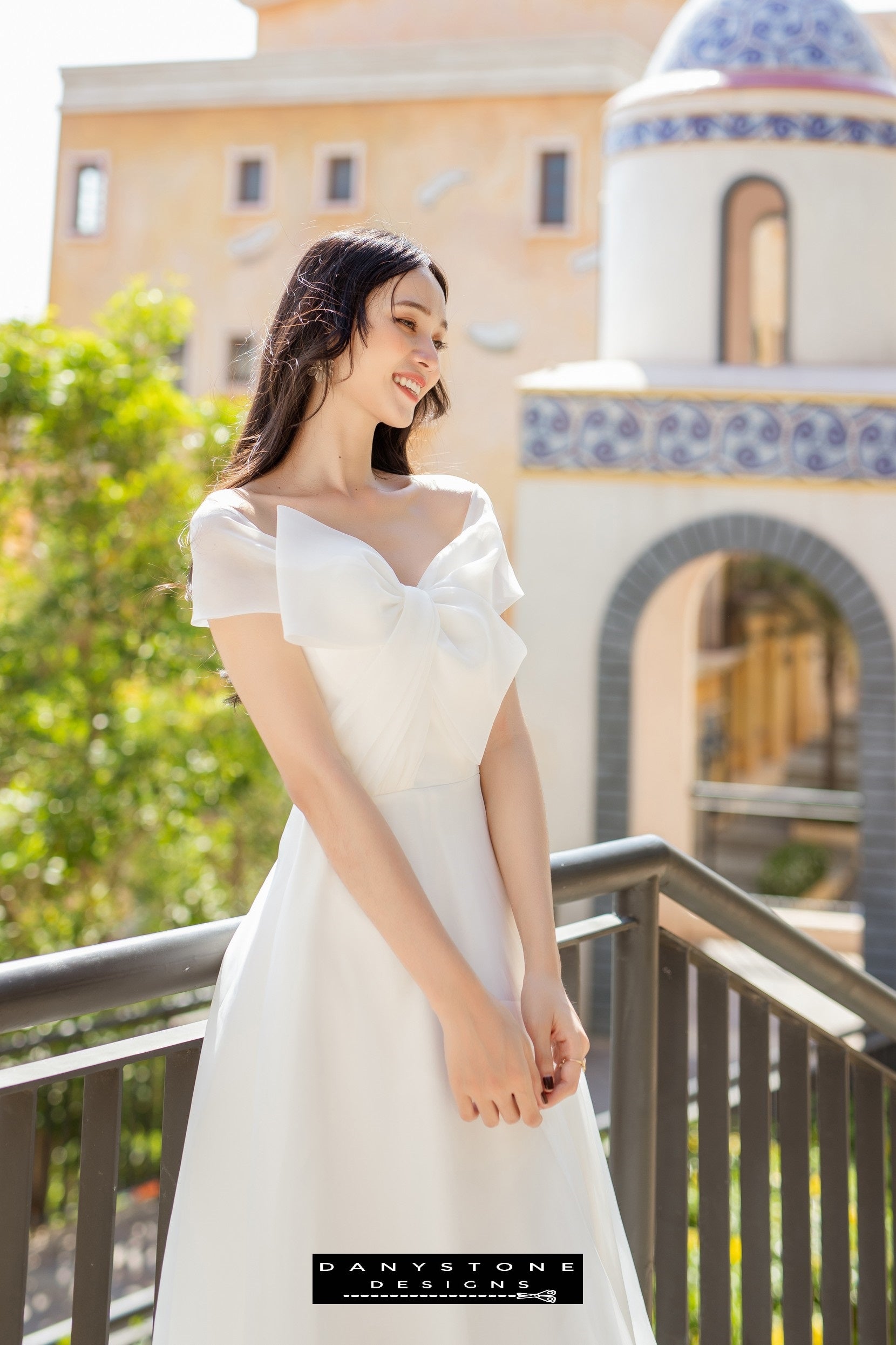 Image 1: Bride wearing a silk chiffon wedding dress with bow accents, smiling on a balcony.