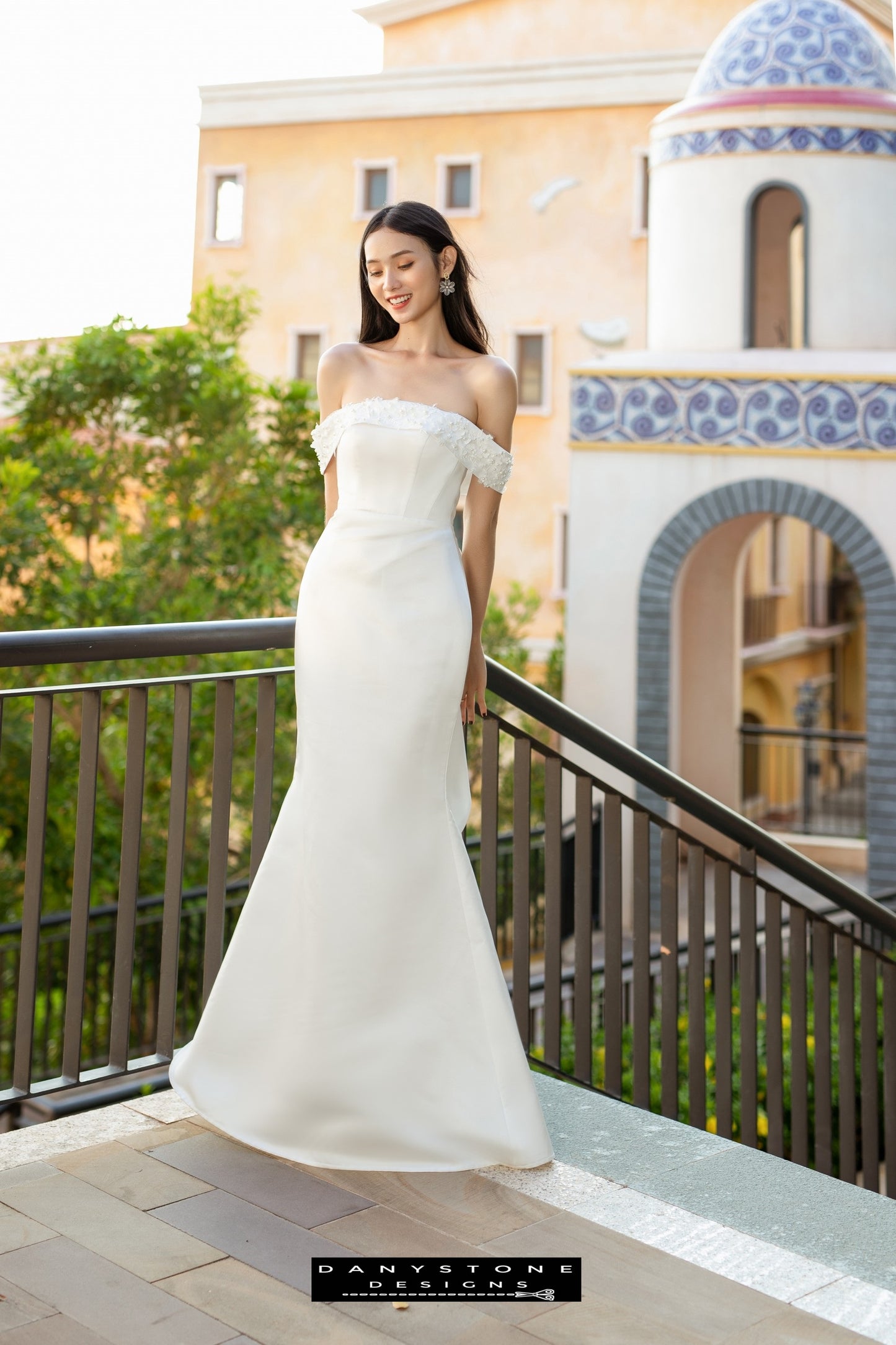 Image 1: Bride wearing a flat shoulder fishtail wedding dress with 3D floral accents, smiling on a balcony.