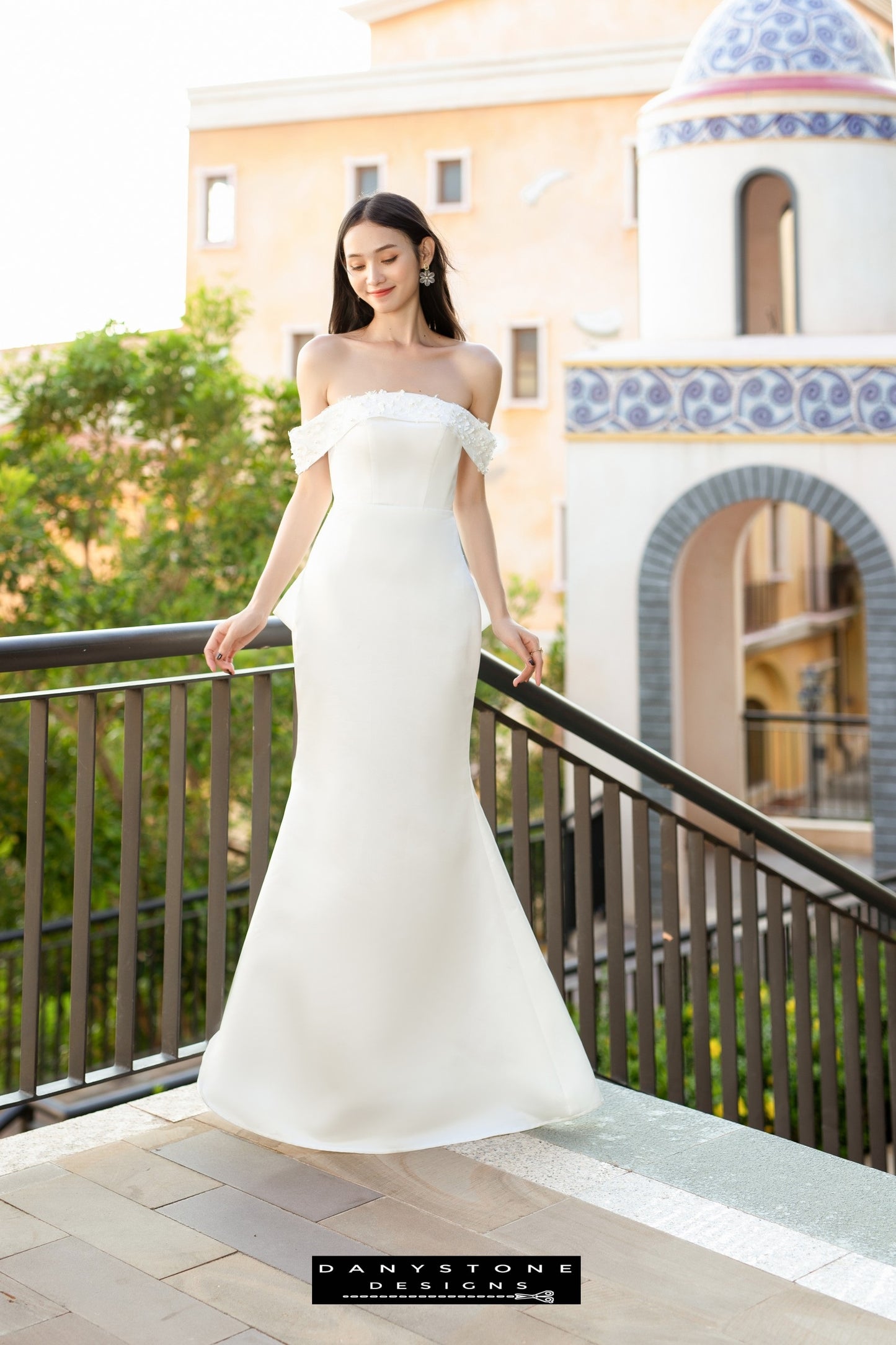 Image 2: Full-length view of a bride in a flat shoulder fishtail wedding dress with 3D floral accents, standing on a balcony.