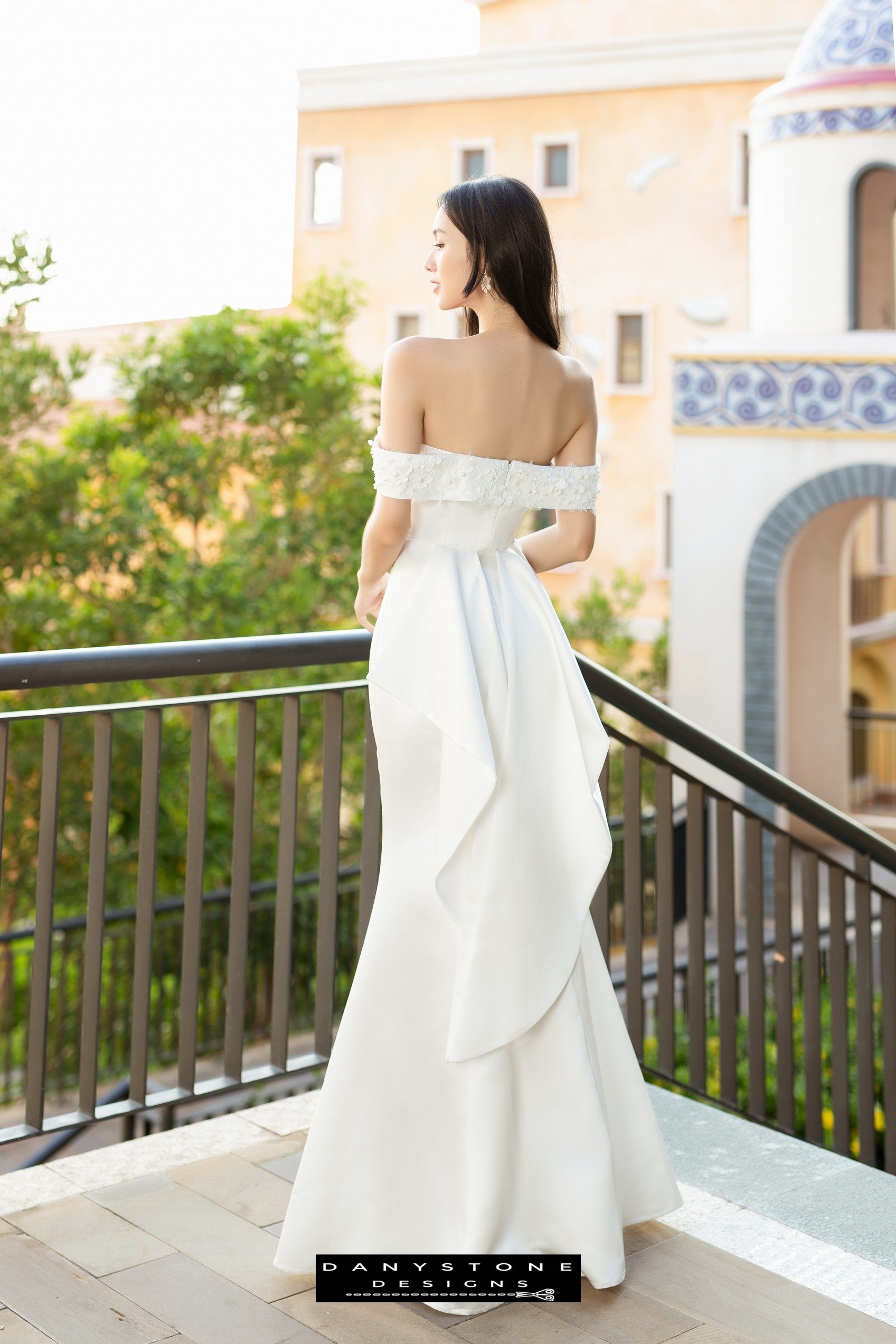 Back view of a bride in a flat shoulder fishtail wedding dress with 3D floral accents, showing the flowing fishtail design.