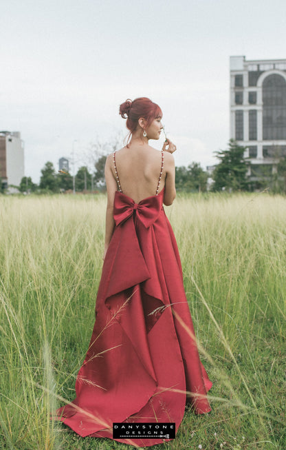 Back view of woman wearing a red evening dress with pearl straps and a large bow, standing in a grassy field