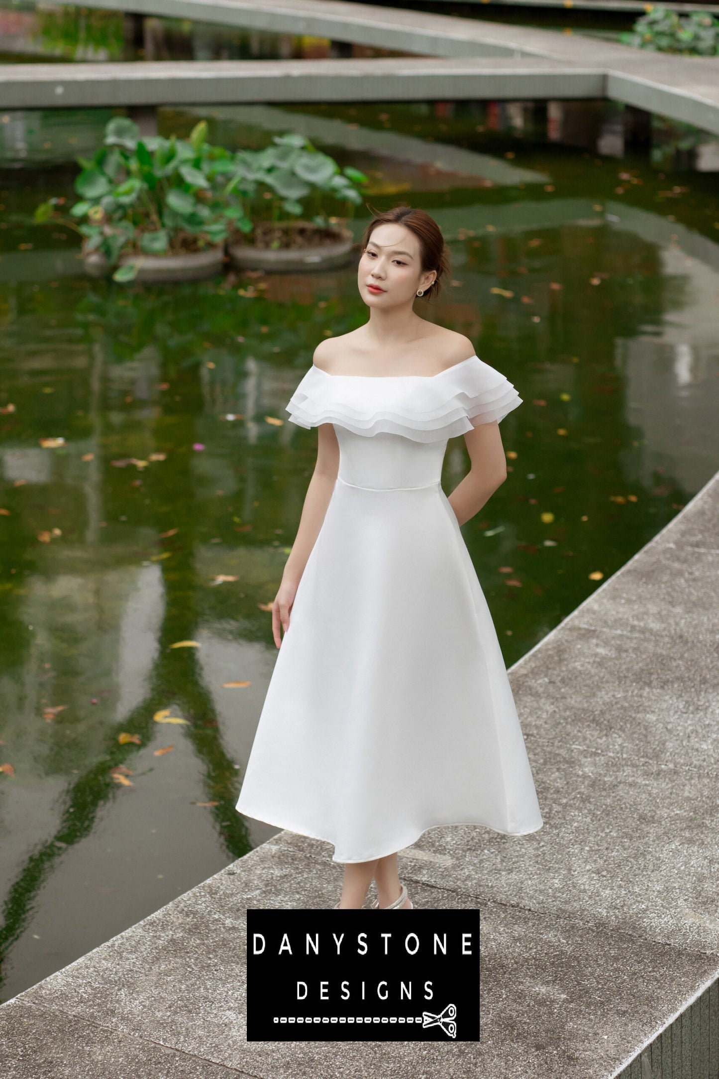 Full view of a woman in a white midi 4-layer flat-shoulder dress, standing on a path by a pond.