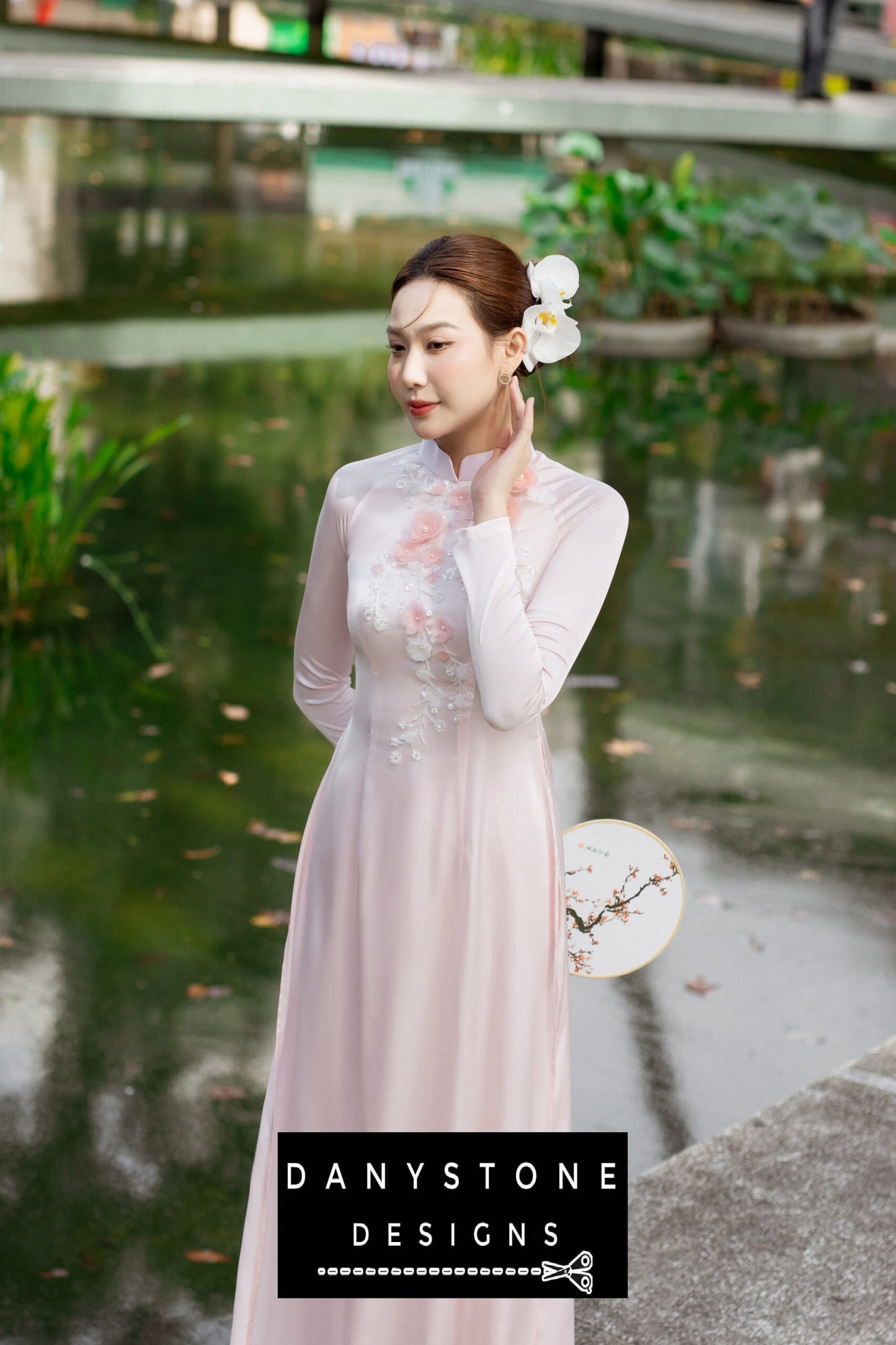 Full-length view of a woman in a pink silk chiffon Áo Dài, standing near a serene pond.