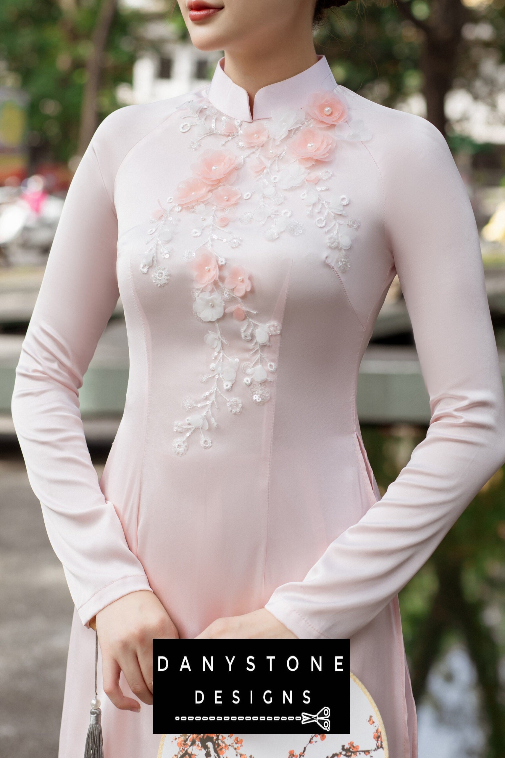 Elegant woman in a pink silk chiffon Áo Dài with floral embellishments, holding a fan.