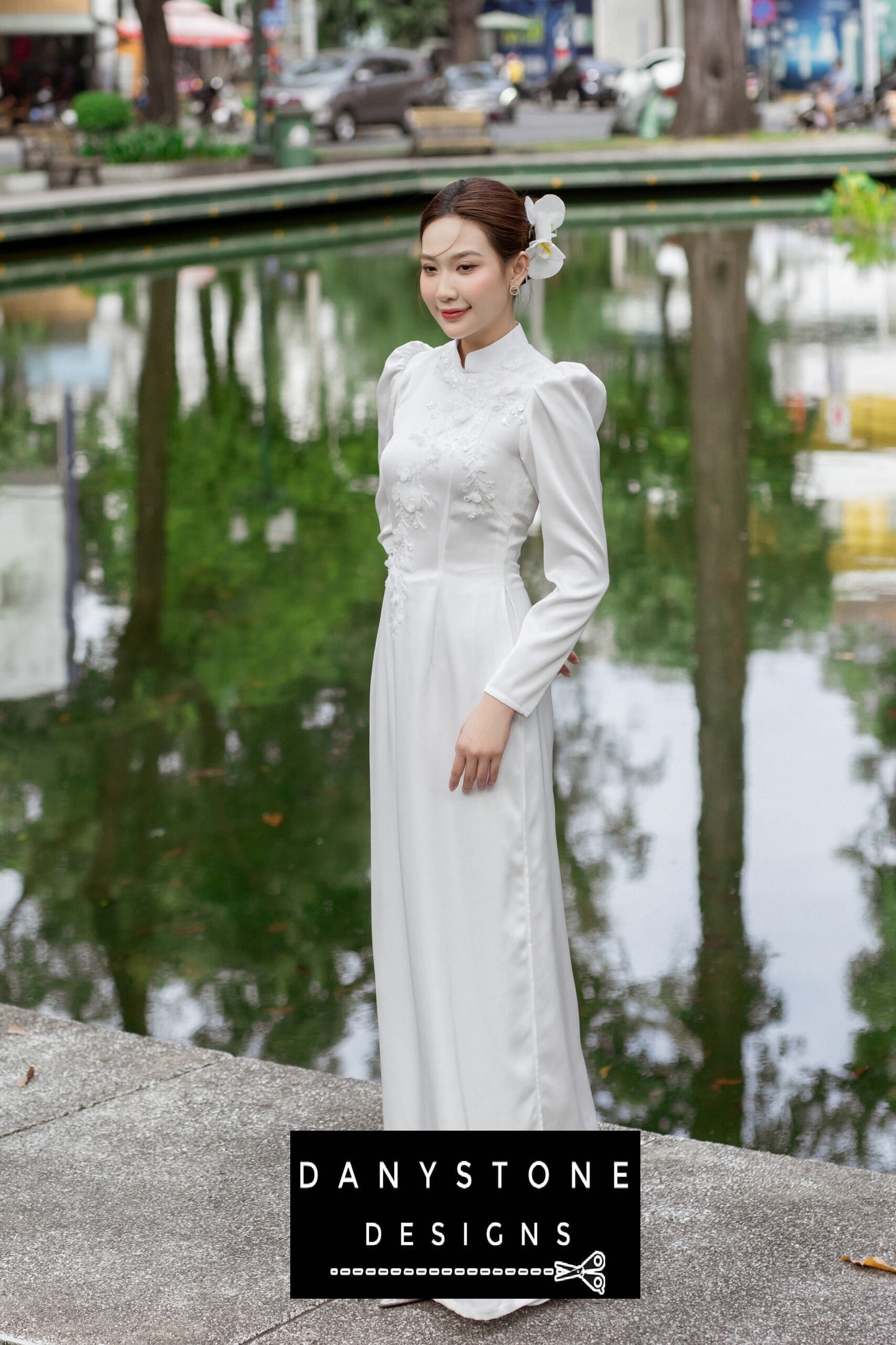 Full-length view of a woman wearing a white silk chiffon Áo Dài, standing by a pond.