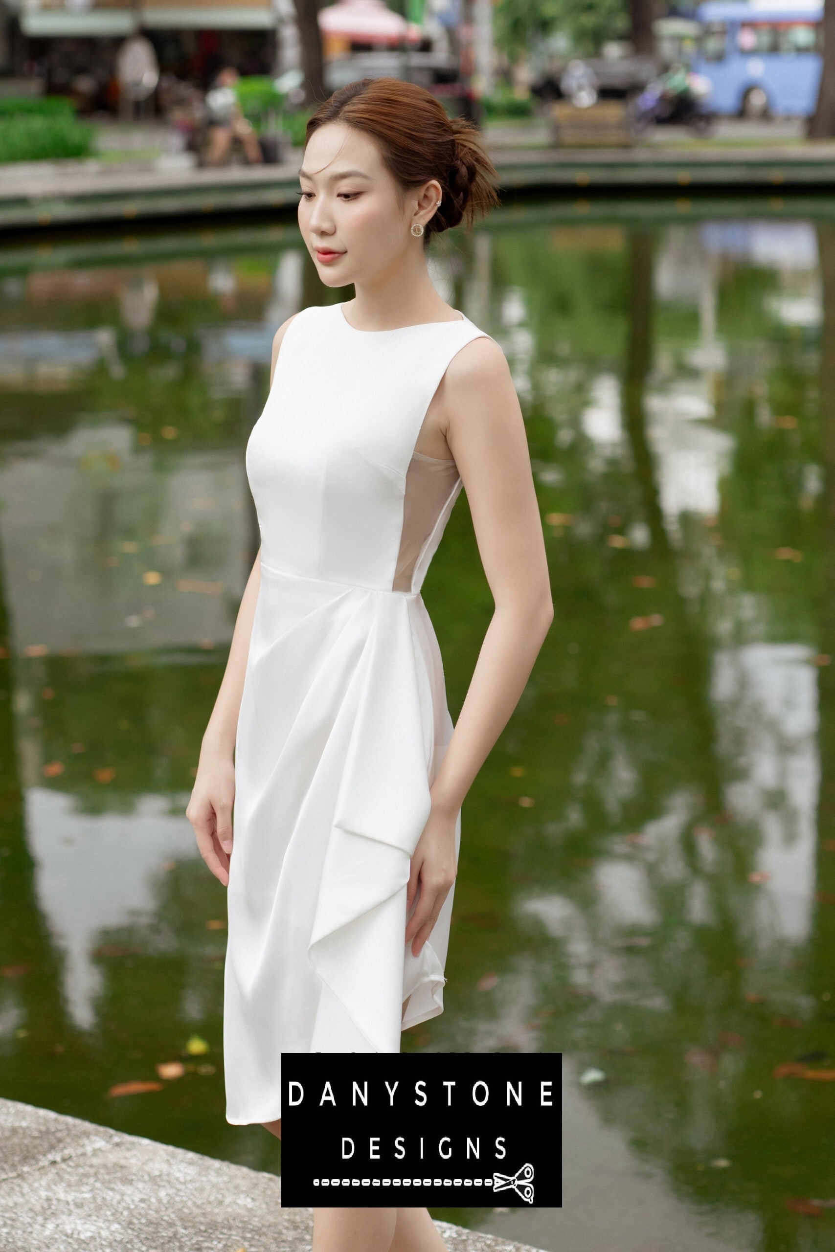 Woman in a short boat neck dress with pleated waist, standing by a serene pond.