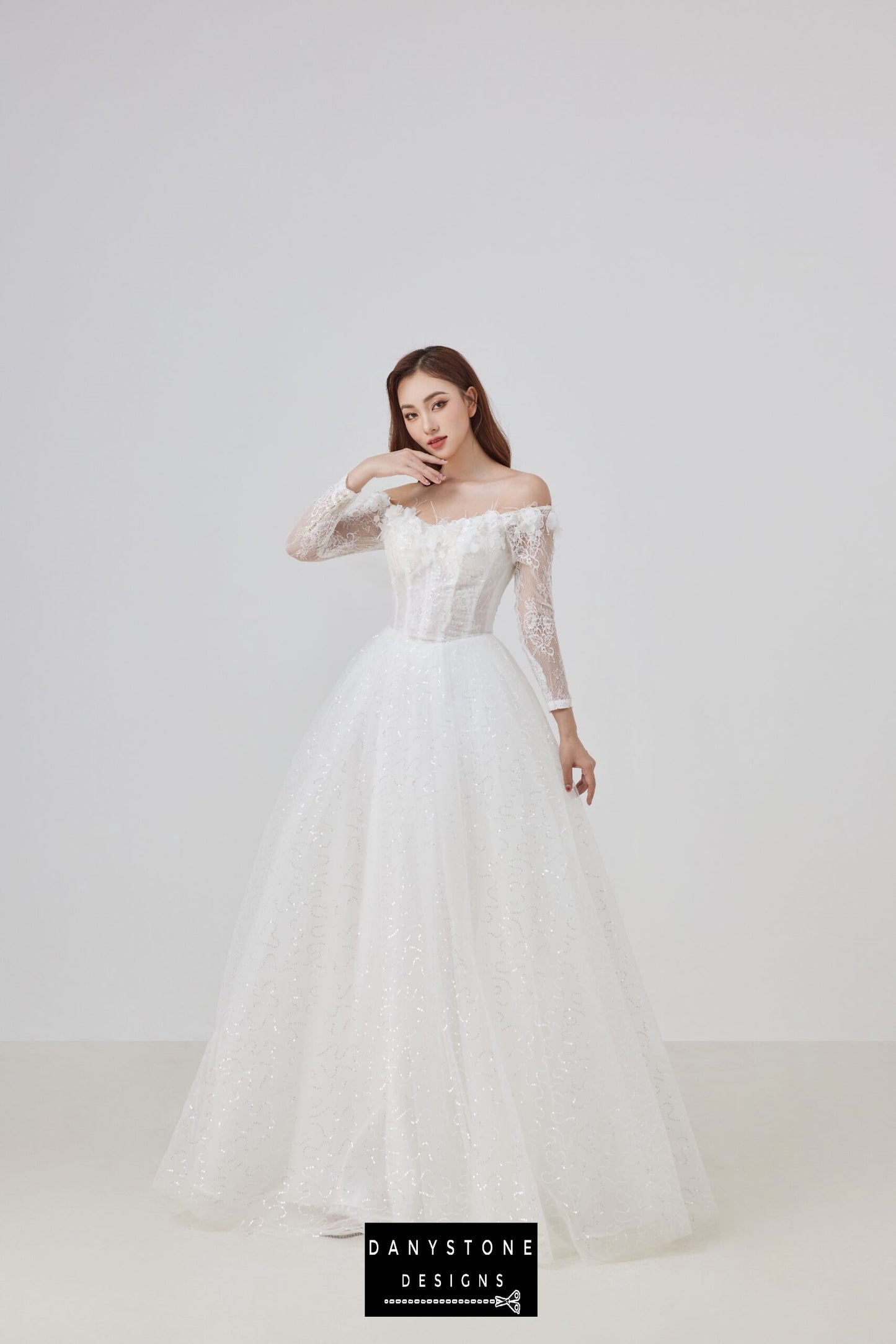 Bride wearing an off-the-shoulder lace wedding dress with 3D flower embellishments, standing with one hand on her chin in a bright studio.