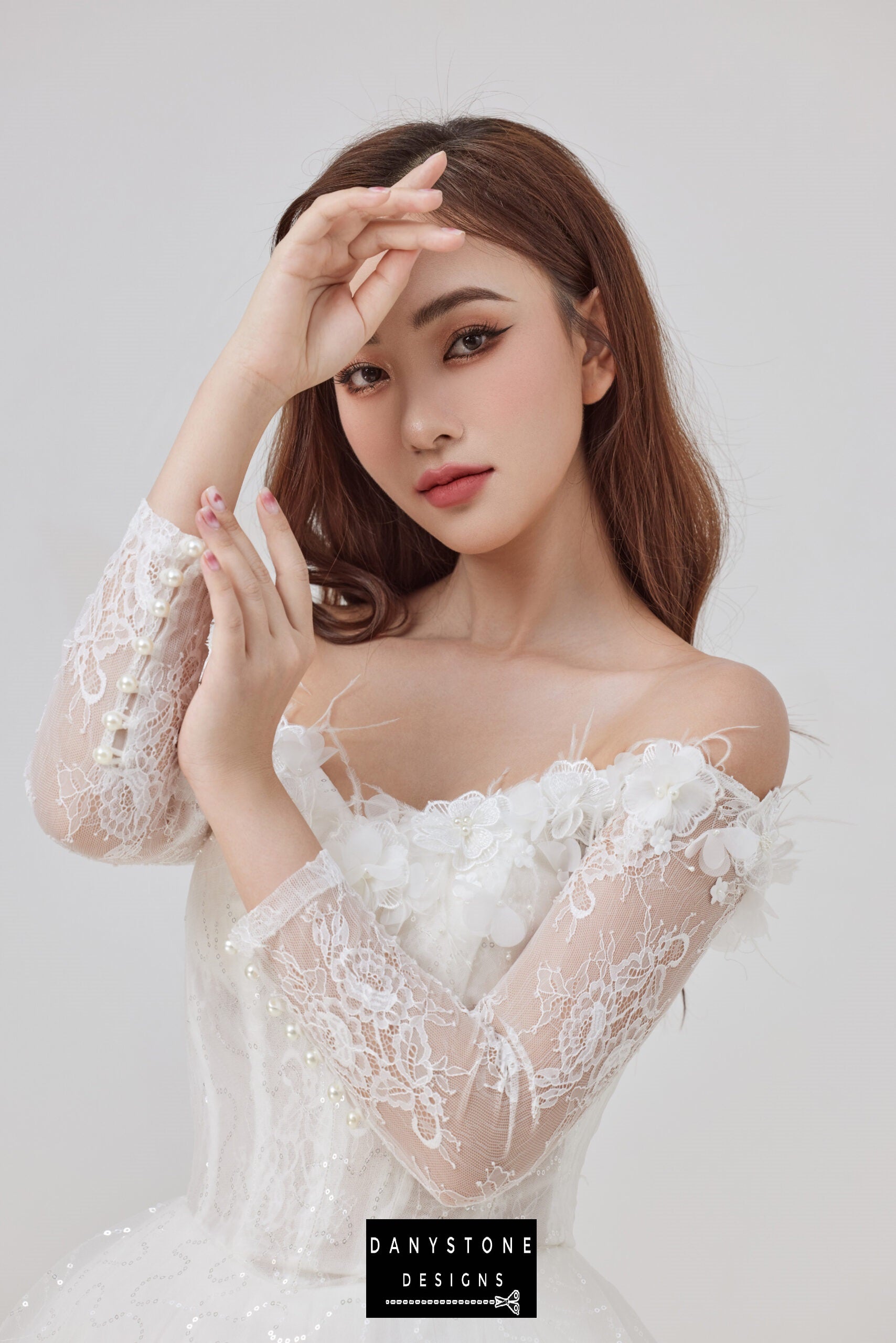 Bride wearing an elegant off-the-shoulder wedding dress with lace sleeves and 3D flower embellishments, posing with one hand raised to her forehead.