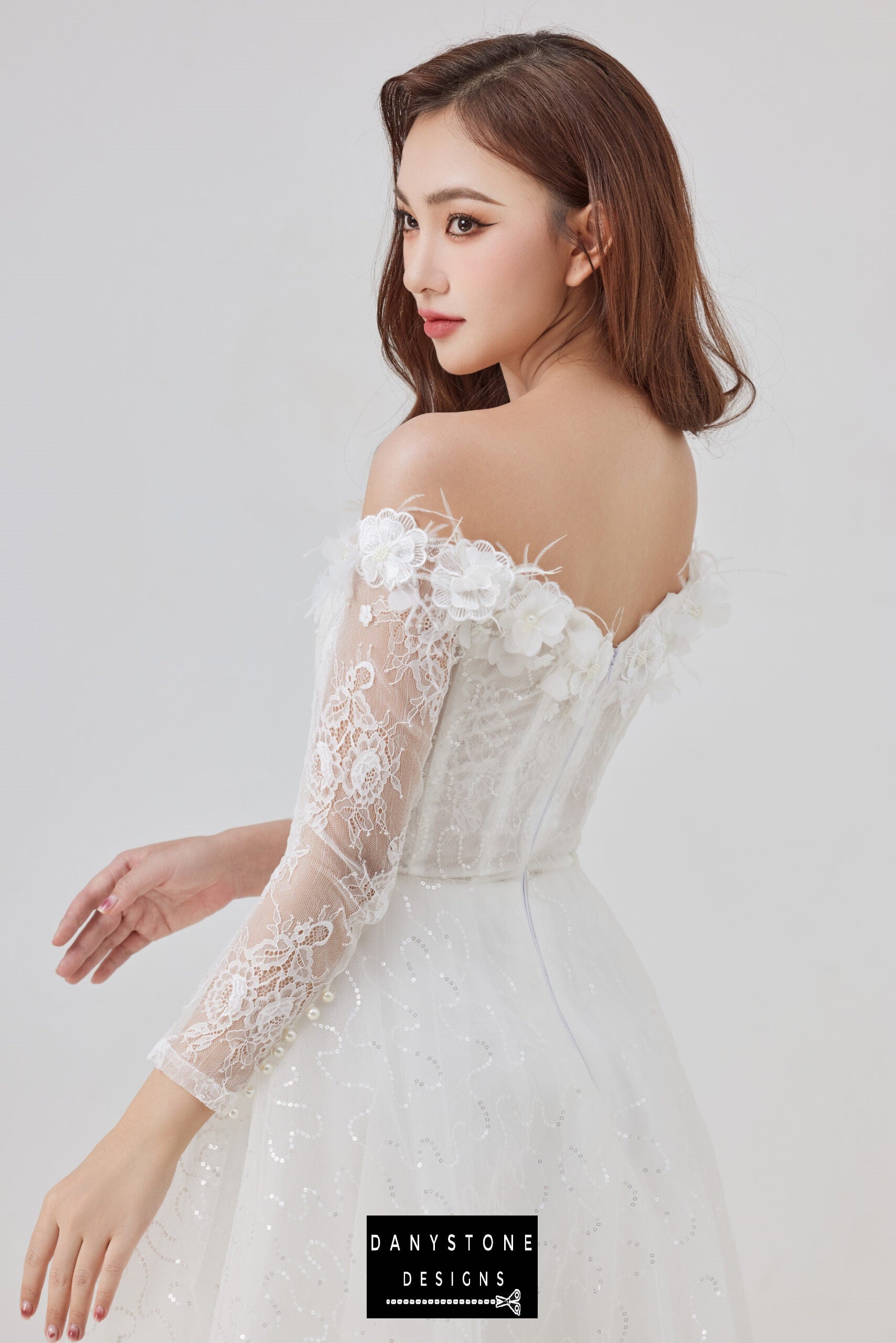 Bride in an off-the-shoulder wedding dress with lace sleeves and 3D flower details, posing with one hand on her hip and looking over her shoulder.