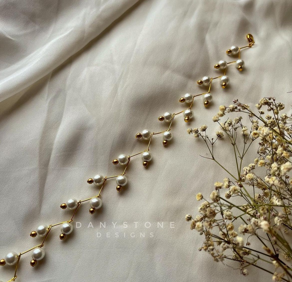 Handmade pearl choker necklace with delicate pearls on a gold-toned chain, displayed on a soft beige fabric background alongside dried flowers.